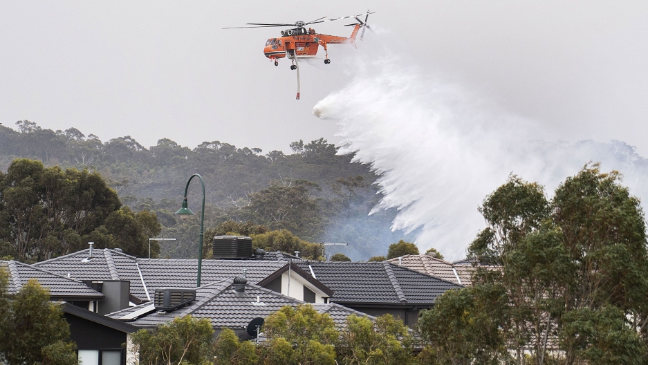 A helicopter drops water on a bushfire behind houses in Bundoora, Melbourne, Australia, Dec. 30, 2019. A large emergency helicopter is shown flying above several houses and dropping a large quantity of water.