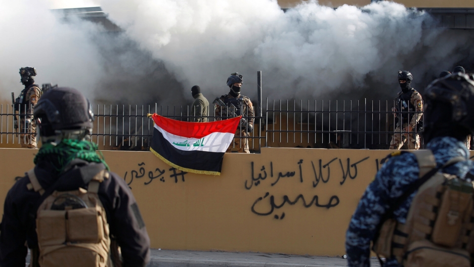 Members of Iraqi security forces are seen in front of US Embassy during a protest to condemn air strikes on bases belonging to Hashd al-Shaabi paramilitary forces, in Baghdad, Iraq, Jan. 1, 2020. Several Iraqi security authorities are shown standing guard behind a metal fence with smoke billowing out from the US Embassy behind them.