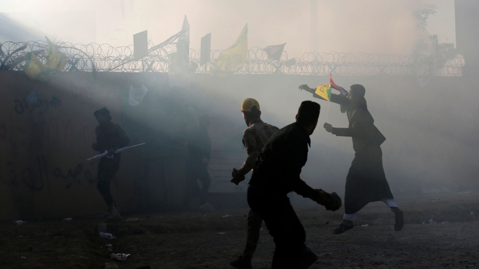 Iran-backed militia fighters throw stones toward the US Embassy during a protest to condemn air strikes on bases belonging to Hashd al-Shaabi paramilitary forces, in Baghdad, Iraq, Dec. 31, 2019. Several people are shown outside of the US Embassy in Baghdad with rocks and other objects in their hands and smoke surrounding the scene.