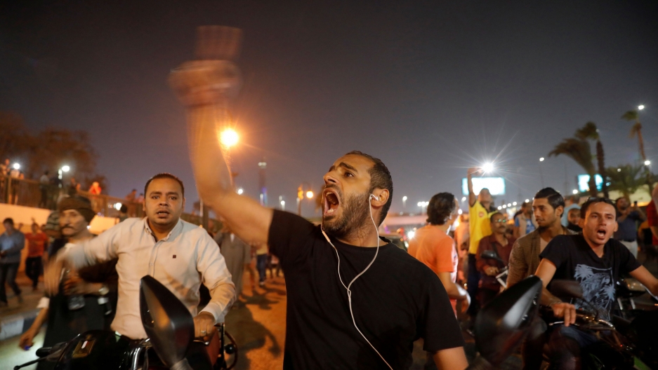 A group of people are shown protesting with one man in the middle with his hand raised and finger pointing to the sky.