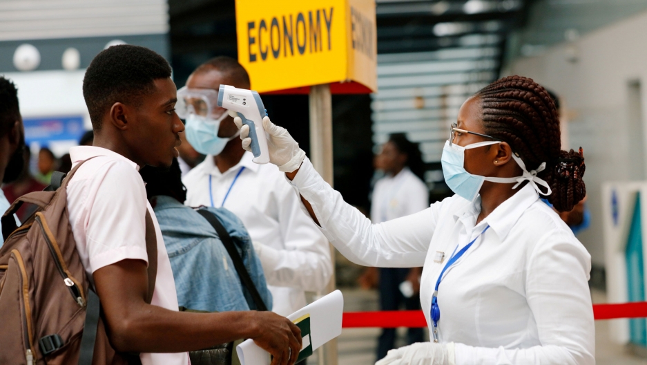 A health worker checks the temperature of a traveler as part of the coronavirus screening procedure at the Kotoka International Airport in Accra, Ghana, Jan. 30, 2020. A woman is shown with a face mask on and holding a device up to the face of a young man opposite her.