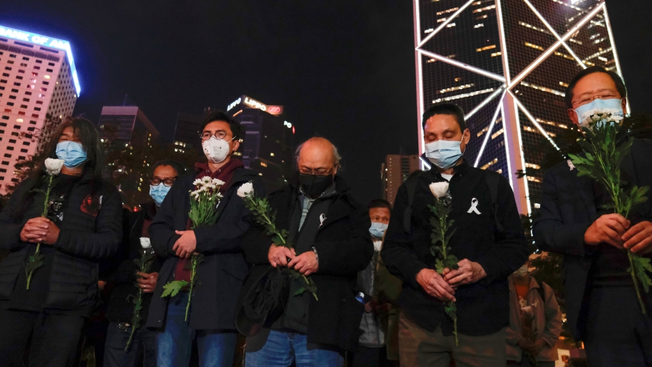 People wear masks as they attend a vigil in Hong Kong for late the Li Wenliang, an ophthalmologist who died of coronavirus at a hospital in Wuhan, China, Feb. 7, 2020. A group of people are shown holding flowers and wearing face masks with skyscrapers in the distance.