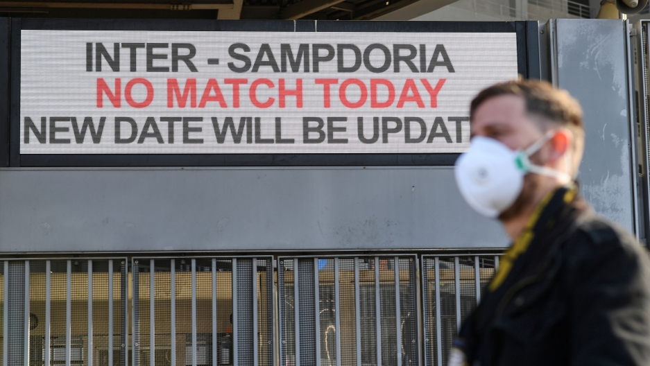A man wearing a face mask stands outside the San Siro stadium after the Inter Milan vs Sampdoria Serie A match was canceled due to an outbreak of the coronavirus in Lombardy and Veneto, in Milan, Italy, Feb. 23, 2020. A man is shown in soft focus wearing a face mask and standing outside the San Siro stadium with a sign that reads, "No Match Today."
