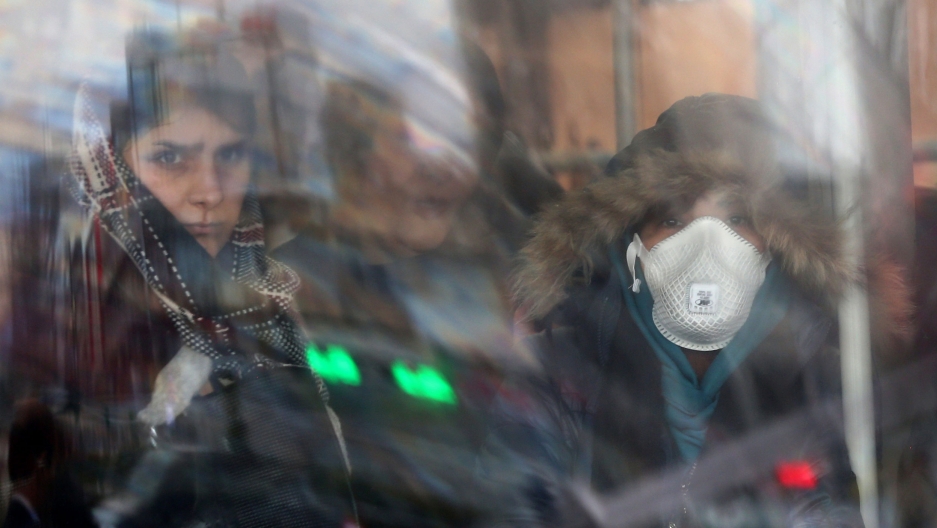 An Iranian woman wears a protective mask as she sits in a bus in Tehran, Iran, Feb. 25, 2020. Women are shown through reflecting glass with one wearing a face mask and a hooded jacket.