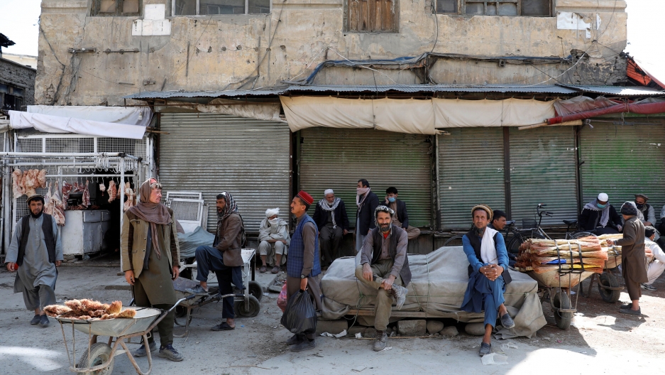 Afghan workers sit as a market is closed during the coronavirus outbreak in Kabul, Afghanistan, April 23, 2020. Several men are shown sitting on a table covered by a cloth tarp with closed market stalls behind them.