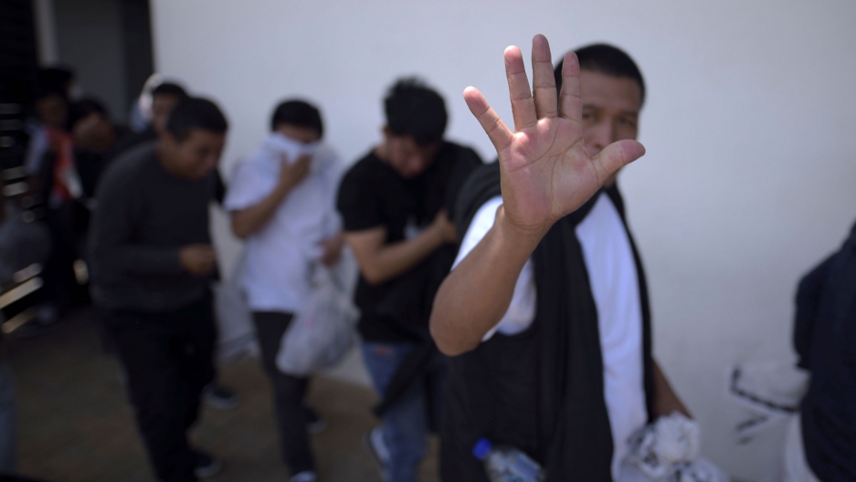 Guatemalan migrants walk toward a bus after being deported from the US outside La Aurora International Airport in Guatemala City, Guatemala, March 19, 2020. A man is show holding his hand up to the camera as he walks with several over people.