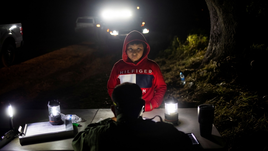 Juan, an 8-year-old unaccompanied minor from Guatemala, is registered by a border patrol agent in a field. A young boy is shown wearing a red hooded sweatshirt and standing at a border patrol table.