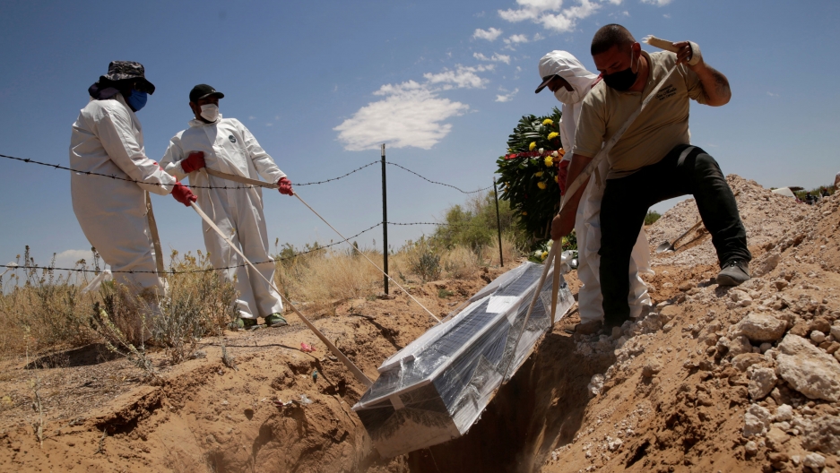 Cemetery workers place a coffin, containing the body of a 52-year old woman who died of the coronavirus disease, into a grave at the San Rafael cemetery, in Ciudad Juarez, Mexico, May 19, 2020. For men are shown wearing protective clothing and lowering a casket wrapped in clear plastic into a grave.