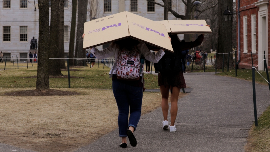 Students carry boxes to their dorms at Harvard University, after the school asked its students not to return to campus after Spring Break to counter the coronavirus outbreak, in Cambridge, Massachusetts, March 10, 2020. Two women are shown carrying cardboard FedEx boxes over their heads as they walk through Harvard University's campus.