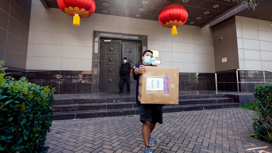 A FedEx employee removes a box from the Chinese Consulate Thursday, July 23, 2020, in Houston. Texas. US has ordered China to close its consulate in Houston. A man is shown wearing a face mask and carrying a large brown cardboard box with the doors of the Chinese Consulate in Houston in the background.