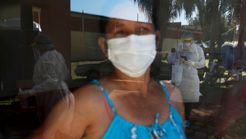A woman in quarantine looks through the window to watch members of the National Mechanism to Prevent Torture visiting a school turned into a government-run shelter where citizens returning home are required by law to quarantine for two weeks and COVID-19 A woman is shown with a face mask and wearing a blue top and looking through glass where several people are shown wearing full medical protective gowns.