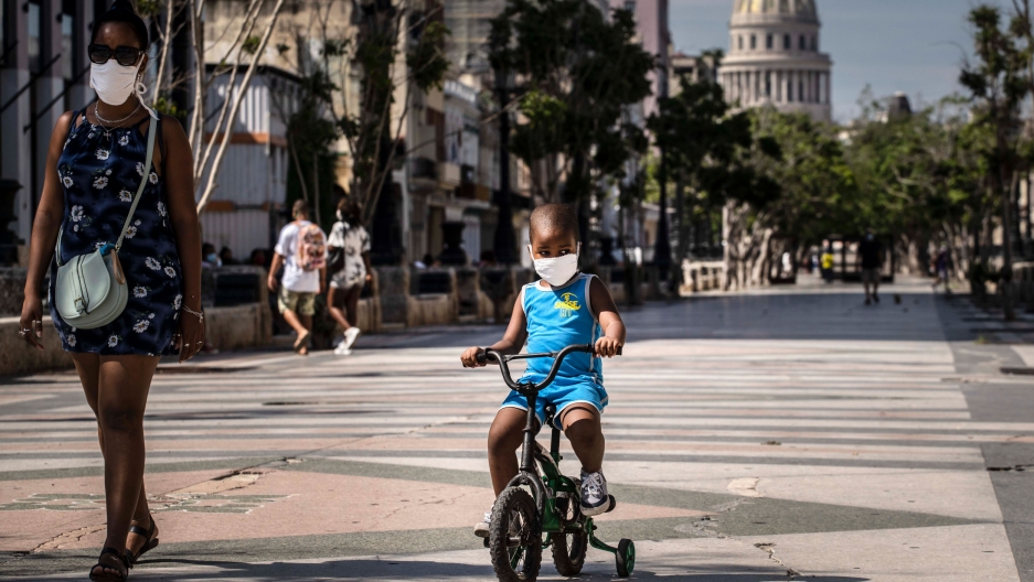 A woman accompanies a child on a bicycle, both wearing protective face masks as a precaution amid the coronavirus pandemic, in Havana, Cuba, Friday, July 3, 2020. A young child is shown wearing a face mask and a blue outfit and riding a small bicycle with training wheels.