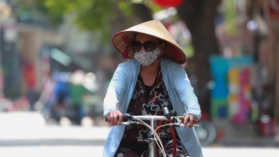 A woman wearing face mask rides a bicycle in Hanoi, Vietnam on Thursday, July 30, 2020. A woman is shown wearing a blue jacket, sunglasses, face mask and a straw hat while writing a bicycle.