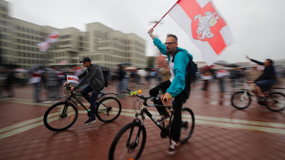 Belarusian opposition supporters ride bicycles and wave an old Belarusian national flags during a protest rally in front of the government building at Independent Square in Minsk, Belarus, Wednesday, Aug. 19, 2020. Several people are shown riding bicycles with one carrying an old Belarusian flag in a photograph with blured motion.