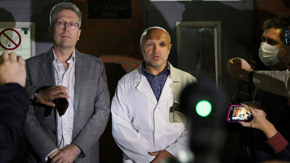 Anatoly Kalinichenko, deputy chief doctor of the Omsk Ambulance Hospital No. 1 (R) and Boris Teplykh, head of the department of anesthesiology and resuscitation of the Pirogov's medical center, speak to the media at the intensive care unit where Alexei Na A man is shown standing with his hands folded and wearing a white medical jacket with members of the media nearby.