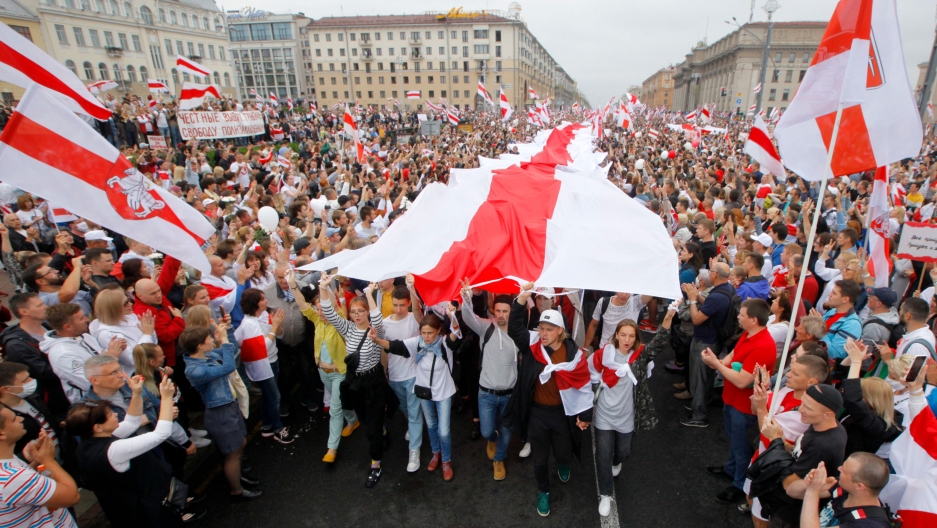 Demonstrators carry a huge historical flag of Belarus as thousands gather for a protest at the Independence Square in Minsk, Belarus, Aug. 23, 2020. A large crowd of people are shown filling Independence Square in Minsk with a long traditional red and white flag streaming through the middle.