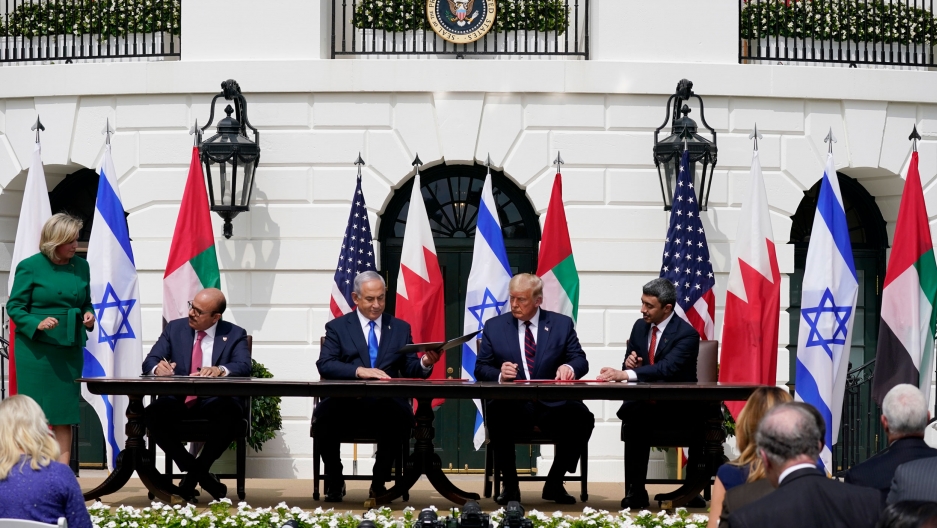 President Donald Trump, center, with from left, Bahrain Foreign Minister Khalid bin Ahmed Al Khalifa, Israeli Prime Minister Benjamin Netanyahu, Trump, and United Arab Emirates Foreign Minister Abdullah bin Zayed al-Nahyan, during the Abraham Accords sign The leaders of the US, UAE, Israel and Bahrain are shown wearing suits and sitting at a table signing documents with flags from each country behind them.