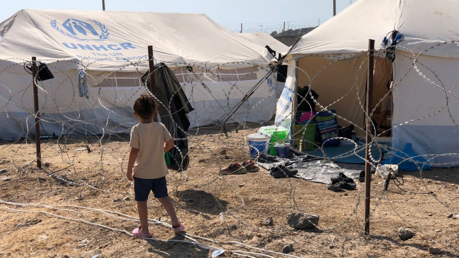 A child walks past new tent shelters in a new refugee camp set up by the Greek government for an estimated 12,000 people displaced by a fire that destroyed the Moria refugee camp on the island of Lesbos. A young child is shown wearing shorts and a t-shirt walking past a barbed-wire fence and white UNHCR tents.