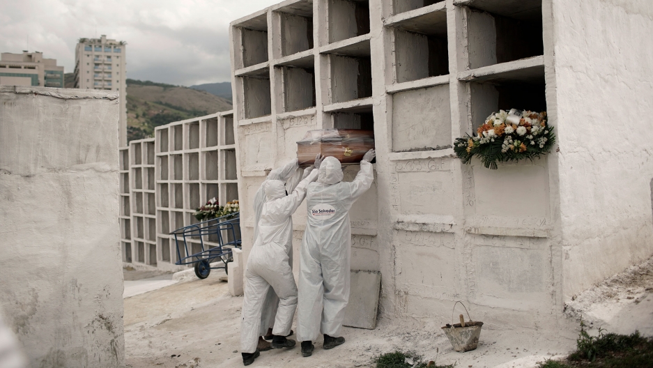 Cemetery workers place the coffin containing the remains of Jose de Arimateia, 65, who died from COVID-19 related complications, into a niche at the municipal cemetery in Nova Iguacu, Brazil, Sept. 24, 2020. Three workers are shown wearing white protective clothing and placing a coffin into an empty slot in large white niche.