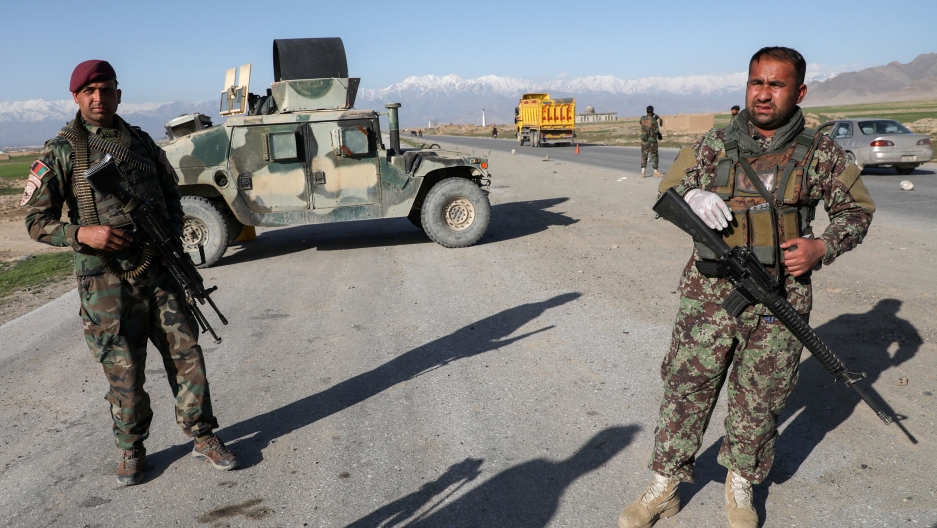 Afghan National Army soldiers stand guard at a check point near the Bagram Airbase, April 2020. Two soldiers are shown standing near a road and holding weapons with an armored vehicle parked behind them.