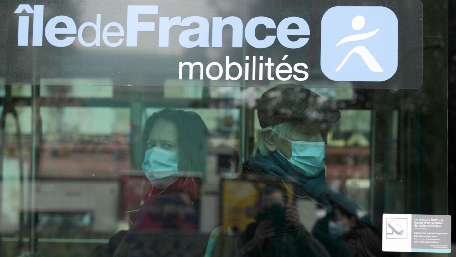 Masked commuters sit in a bus in Paris, Oct. 15, 2020. French President Emmanuel Macron has announced that millions of French citizens in several regions around the country, including in Paris, will have to respect a 9 p.m. curfew starting Saturday. A man and a woman are shown sitting back to back in a bus and looking through a glass window.