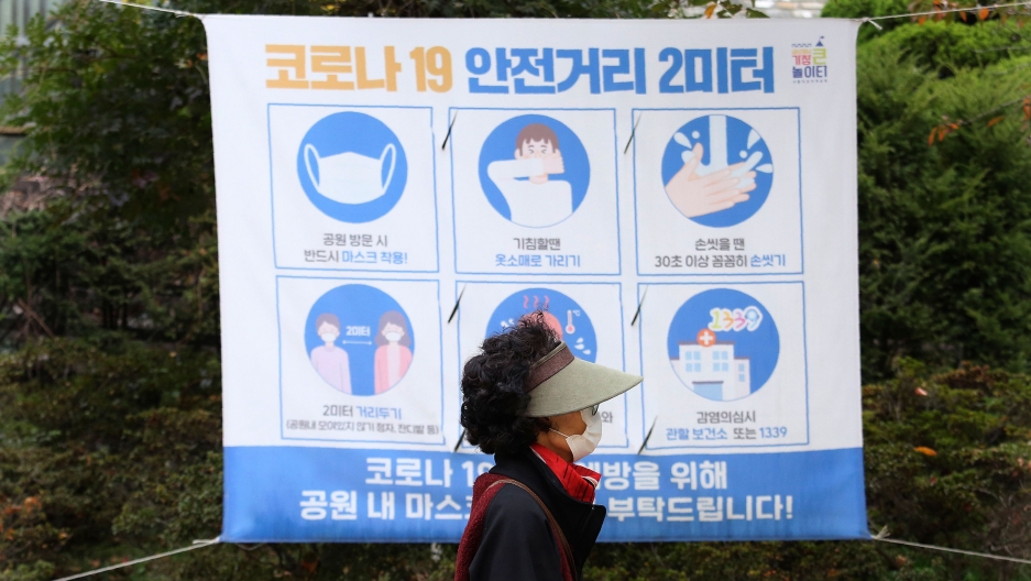 A woman wearing a face mask walks by a social distancing sign at a park in Seoul, South Korea, Oct. 16, 2020. A woman is shown wearing a visor and face mask while walking past a poster with guidance on stopping the spread of the coronavirus.