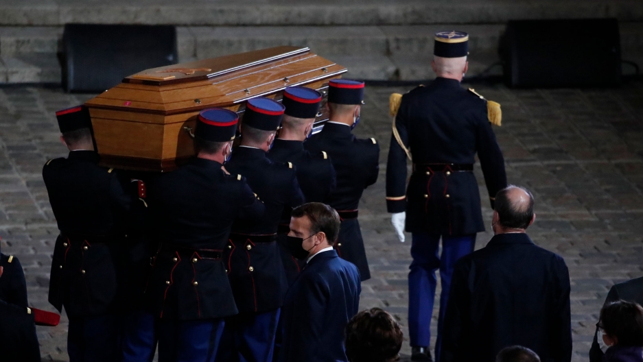 The coffin of slain teacher Samuel Paty is carried in the courtyard of the Sorbonne University during a national memorial event, Oct. 21, 2020, in Paris, France. Eight uniformed officers are shown carrying a brown wooded coffin.