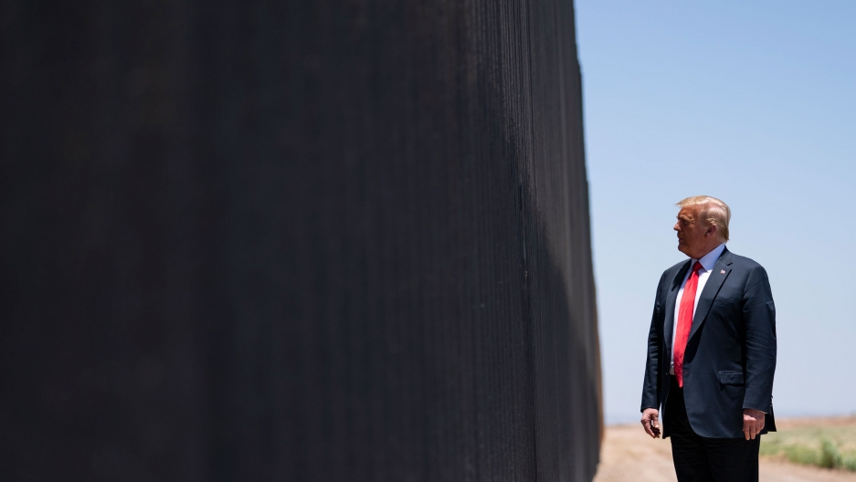 President Donald Trump tours a section of the southern border wall between the US and Mexico in San Luis, Ariz. June 23, 2020. President Donald Trump is shown wearing a blue suit and red tie while looking at the dark facade of the US-Mexico border wall.