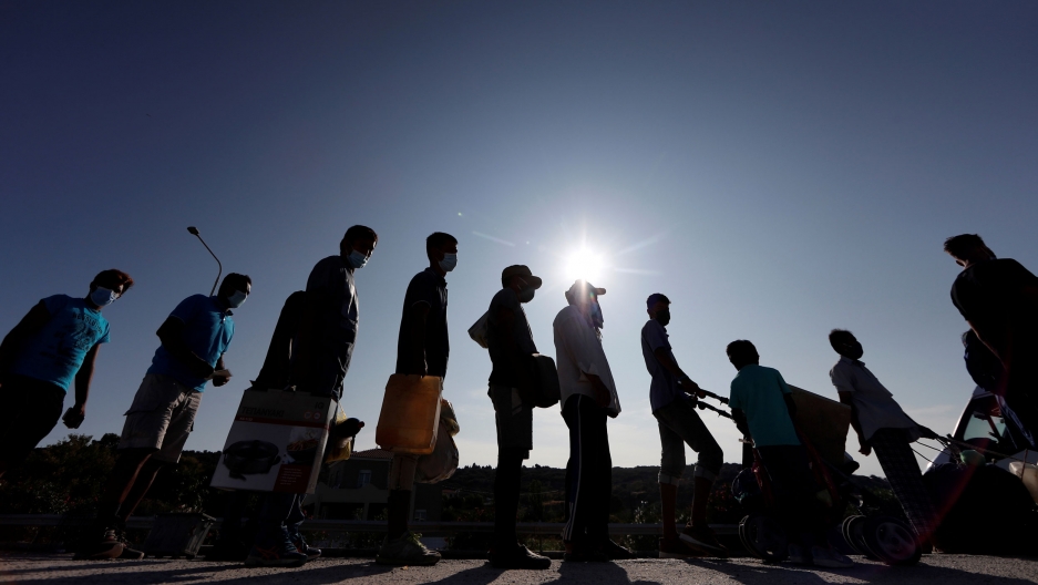 Refugees and migrants line up to enter a temporary camp, on the island of Lesbos, Greece, Sept. 23, 2020. Refugees and migrants are shown in shadow standing in a lin carrying various belongings with the sun flaring in the distance.