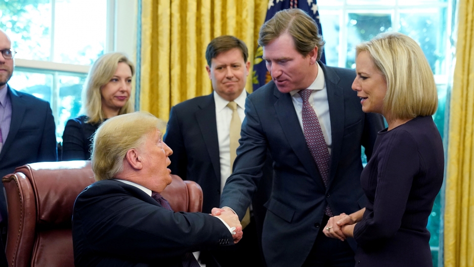 US President Donald Trump shakes hands with Chris Krebs, then director of the Cybersecurity and Infrastructure Security Agency (CISA) as then DHS Secretary Kirstjen Nielsen (R) looks on after a signing ceremony at the White House. Nov. 2018. US President Donald Trump is shown sitting in a large brown chair and leaning over to his left to shake hands with Chris Krebs.