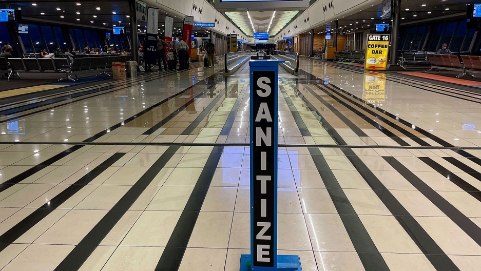 A hand sanitizing pump stands in the middle of a near empty departure terminal as few passengers board and Air France flight to Paris at Johannesburg's airport, Dec. 21 2020. An airport terminal is shown nearly empty with black stripes along the flooring and a large hand sanitizing station in the center of the photogragh.