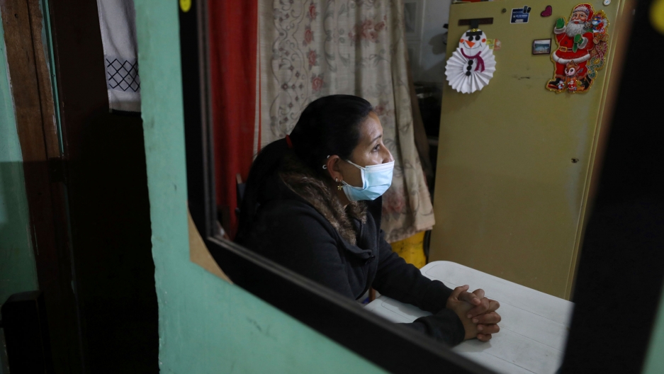 Venezuelan migrant Yaquelin Timaure is seen reflected on a mirror at her home in Bogotá, Colombia, Dec. 21, 2020. A woman is shown seated at a table with her hands crossed and wearing a face mask with a magnet of Santa Claus on the refrigerator in the background.