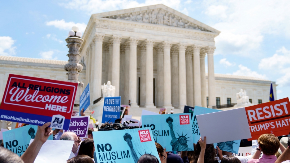 Protesters hold up signs and call out against the US Supreme Court ruling upholding former President Donald Trump's travel ban in Washington, June 2018. A group of people are shown holding different placards protesting the Trump administration's travel ban with the US Supreme Court building in the distance.