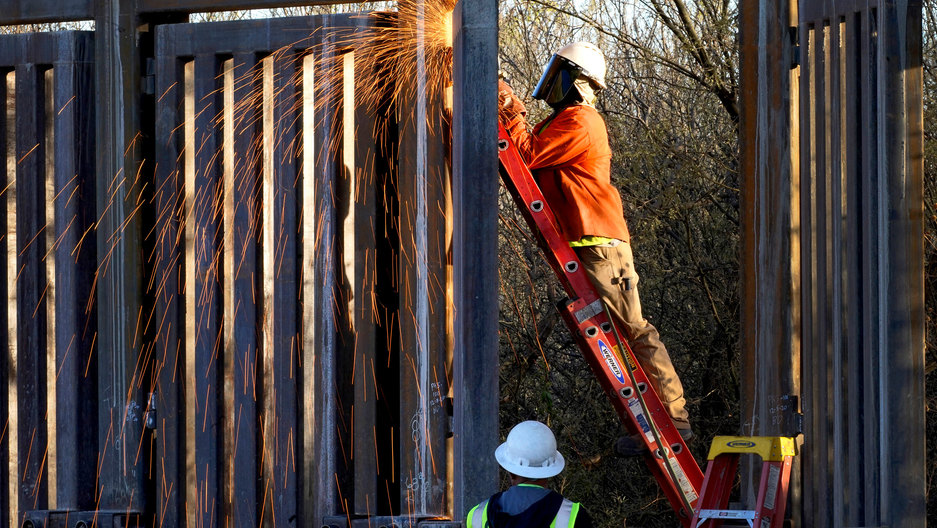 Crews construct a section of border wall in San Bernardino National Wildlife Refuge in Arizona, Dec. 8, 2020. President Biden has ordered a "pause" on all wall construction. A man is shown standing on a ladder and using a machine causing sparks to fly in the construction of a border wall.