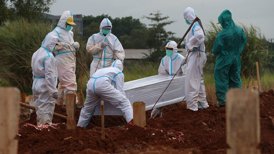 Workers prepare a coffin for burial at the special section of the Padurenan cemetery opened to accommodate the surge in deaths during the coronavirus outbreak in Bekasi, Indonesia, Jan. 28, 2021. Several people are shown in white medical outfits from head-to-tow with to of them lowering a casket into the ground.