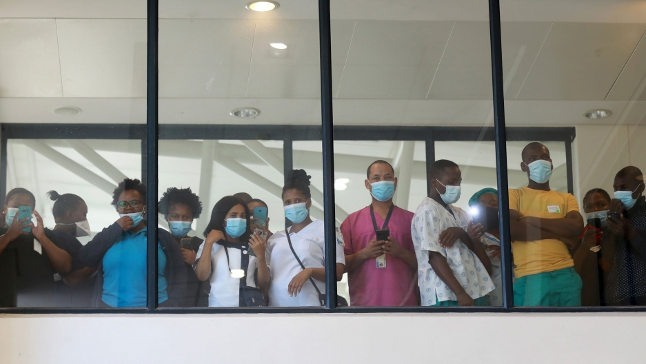 Health care workers look through a window at a hospital in Cape Town, South Africa, during the roll out of the first batch of Johnson and Johnson vaccines in the country. A small crowd of people are shown standing shoulder-to-shoulder, viewed through glass in a photo taken from below.