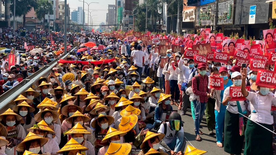Anti-coup protesters fill the main road during a rally in Mandalay, Myanmar, Feb. 22, 2021. A large crowd of people are shown filling a street with many wearing traditional Asian hats and carrying red signs.
