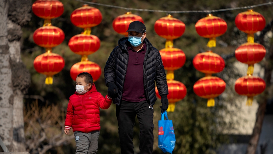 A man and child wearing face masks to protect against the spread of the coronavirus walk past lanterns at a public park in Beijing, Feb. 19, 2021. A man is shown wearing a black puffy jacket while hold the hand of a child wearing a red puffy jacket.