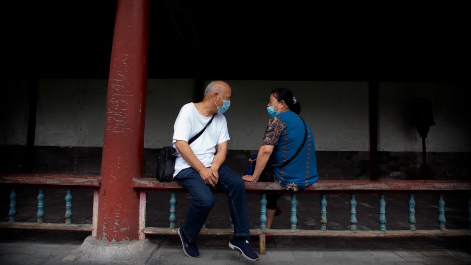 People wearing face masks to protect against the coronavirus sit on a railing at in Beijing, July 2020. A man and woman are shown sitting on either side of a wooden railing and facing each other while wearing protective masks.