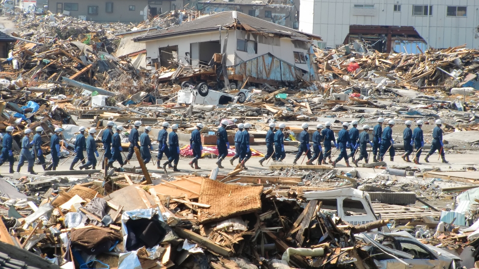 Japan's Self Defense Forces walk through the destruction of Ishinomaki helping aid in the evacuation, 2011. A long line of Japan's Self Defense Forces are shown wearing blue protective outfits and walking among the rubble of Ishinomaki.