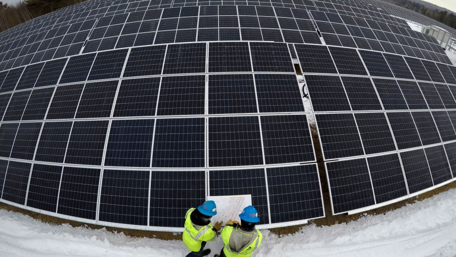 Electricians consult a wiring schematic while installing solar panels at the 38-acre BNRG/Dirigo solar farm in Oxford, Maine, Jan. 14, 2021. Several rows of large solar panels are shown with a wide angle lens with two people at the bottom of the photo wearing yellow safety vests and hard hats.