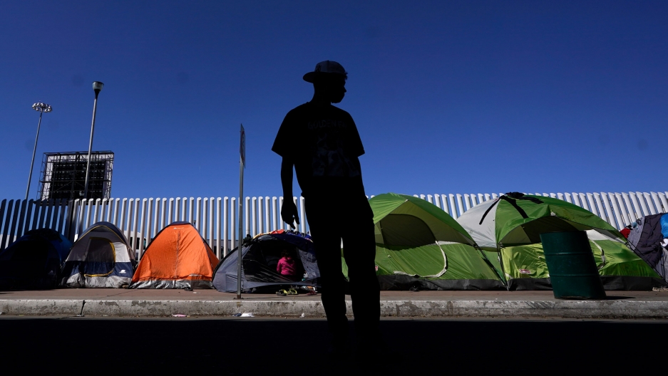 A migrant from Honduras seeking asylum in the United States stands in front of rows of tents at the border crossing in Tijuana, Mexico, March 1, 2021. A man is shown in shadow and wearing a brimmed hat with several pup tents shown in focus in the background.