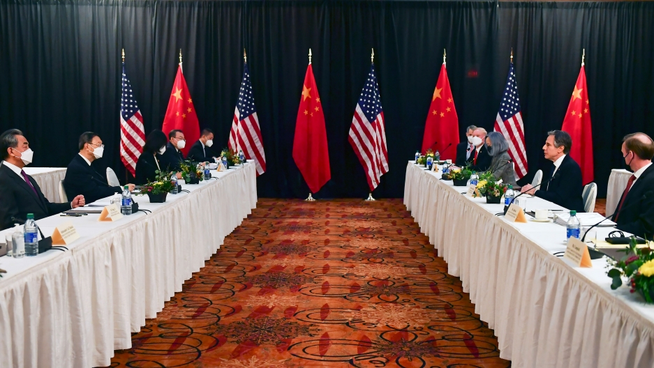 Secretary of State Antony Blinken, on right, speaks during the opening session of US-China talks in Anchorage, Alaska, March 18, 2021. Two long rows of tables are shown with white table cloths and officials on either side with US and Chinese flags in the distance.