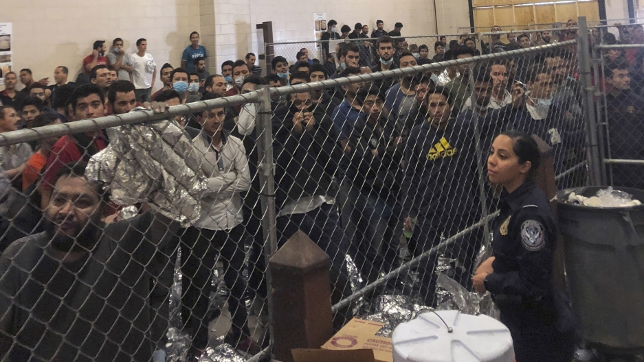 In this July 12, 2019 file photo, men stand in a US Immigration and Border Enforcement detention center in McAllen, Texas, during a visit by Vice President Mike Pence. A group of men stand behind a fenced area within a detention center at the border.
