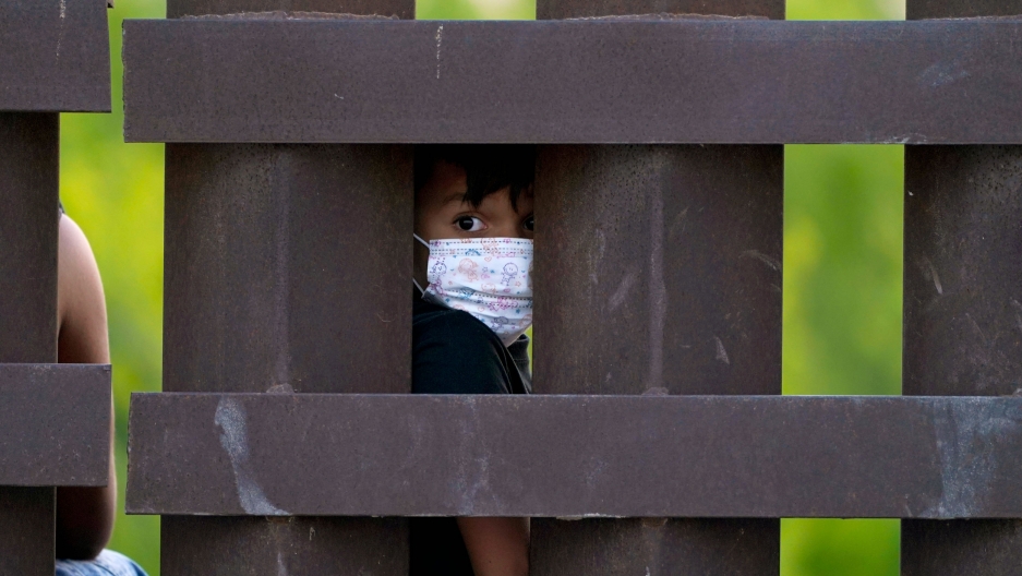 A migrant child looks through the US-Mexico border wall as a group is processed and taken into custody in Abram-Perezville, Texas, March 21, 2021. A small child is shown wearing a protective face mask and looking through the large medal polls of the US-Mexico border fence.