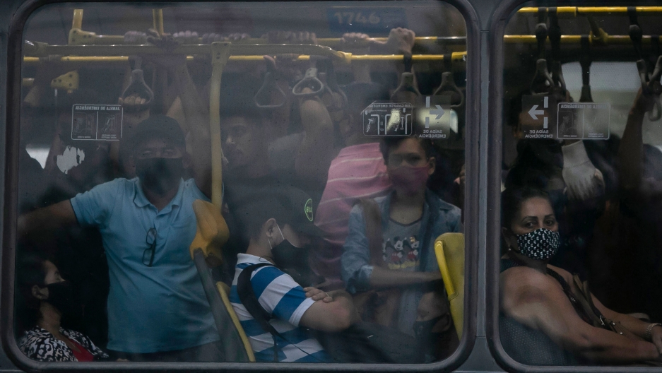 Commuters wearing masks due to the COVID-19 pandemic crowd a public Rapid Transit Bus in Rio de Janeiro, Brazil, March 30, 2021. A crowd of people are shown crammed onto a bus as shown through the window.