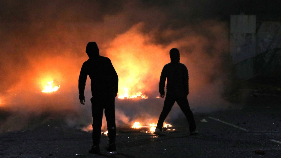 Nationalists and loyalists clash with one another at the peace wall on Lanark Way in West Belfast, Northern Ireland, April 7, 2021. Two people are shown in shadow with a large fire shown in the distance.