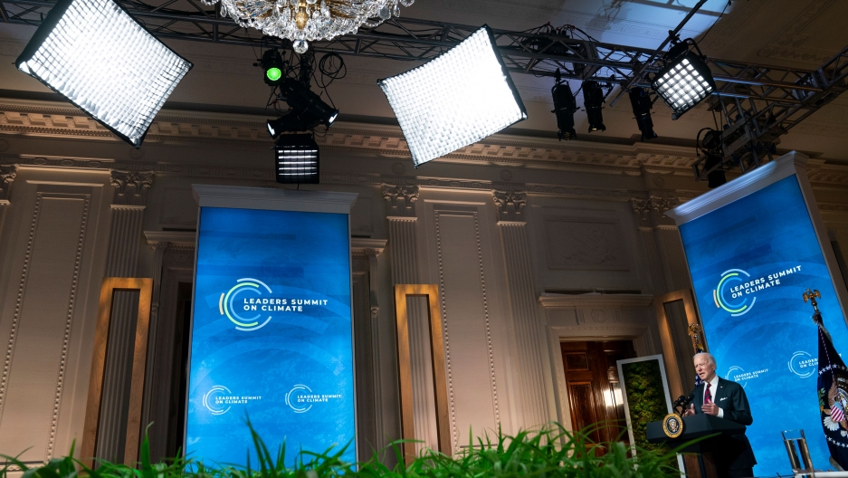 US President Joe Biden speaks to the virtual Leaders Summit on Climate, from the East Room of the White House, April 22, 2021. US President Joe Biden is shown standing at a podium in front of a blue banner in a wide photo also showing the video lighting equipment.