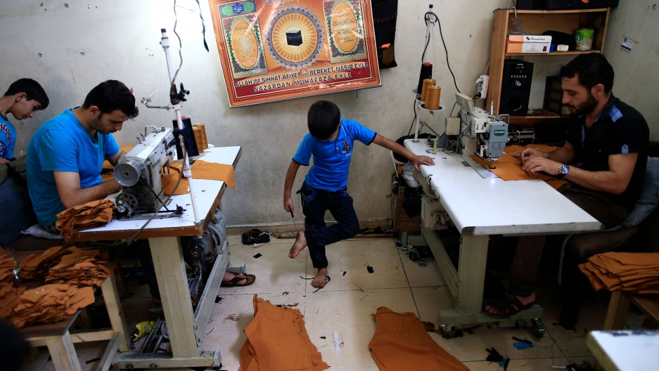 Syrian refugees, including children, work at a clothing workshop in Gaziantep, southeastern Turkey, June 2016. Two men are shown sitting at tables with sewing machines while a young boy is barefoot in the middle.