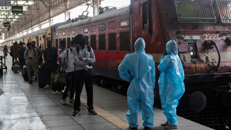 Health workers wait to screen passengers at the Chhatrapati Shivaji Maharaj train Terminus in Mumbai, India, April 15, 2021. A long line of people are shown standing next to a train with two health care workers, shown at the end of the platform, wearing protective clothing.